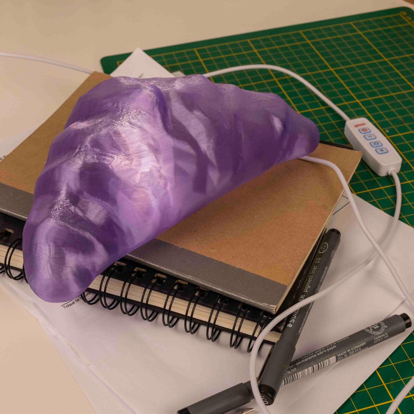Purple croissant lamp on a notebook with pens and a USB cable on a green cutting mat.