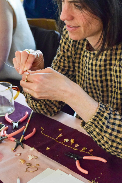 A woman wearing a yellow and black checkered shirt sits at a table, carefully working on jewelry with small gold pieces, pink-handled pliers, and tools on a burgundy runner.